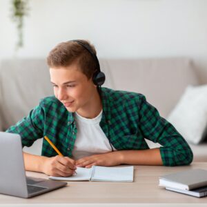 Joyful teen guy writing at notebook and looking at laptop